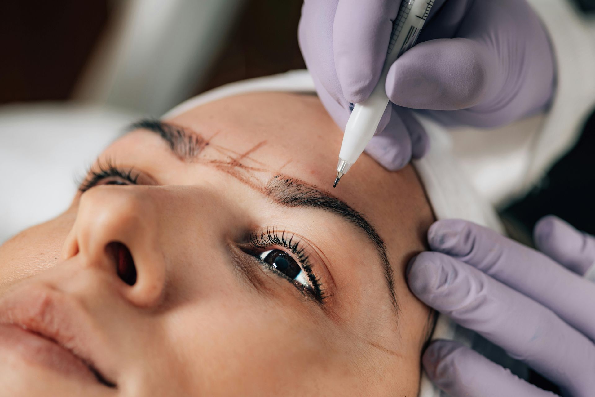 A technician in purple gloves uses a white pen to outline eyebrows for a cosmetic procedure.
