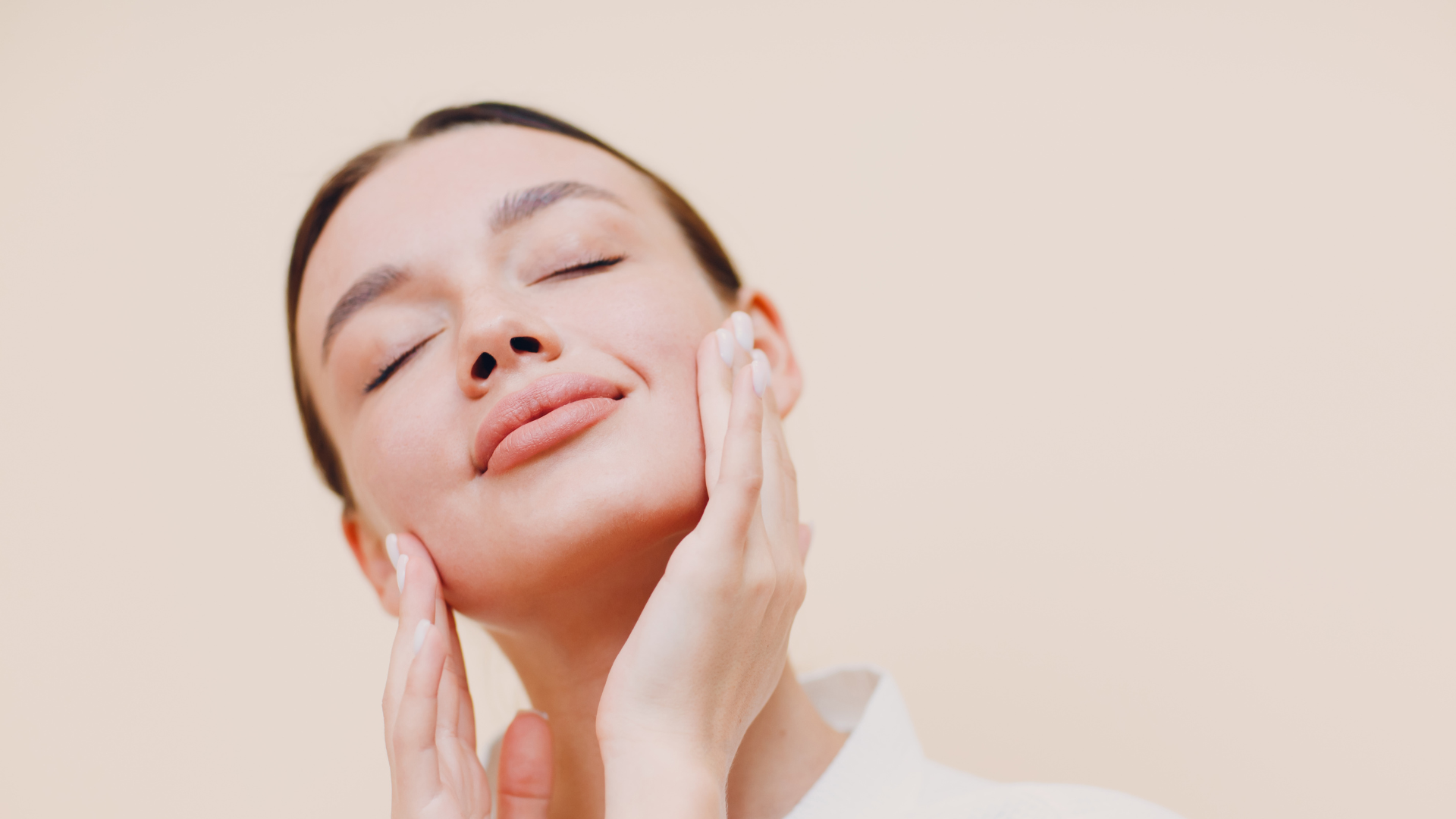 A person with eyes closed gently touches their cheek against a plain beige background.
