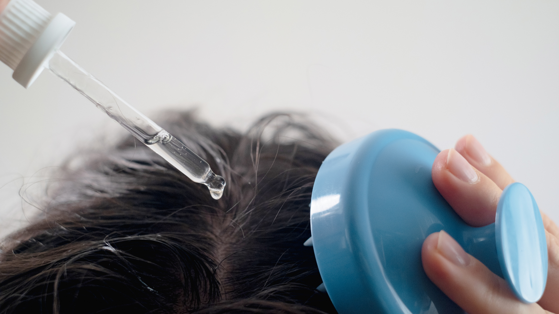 A hand uses a dropper to apply serum to a scalp, while another hand holds a blue scalp massager nearby.