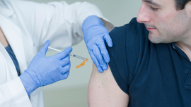 A medical professional in a white coat and blue gloves administers an injection into a person's upper arm.