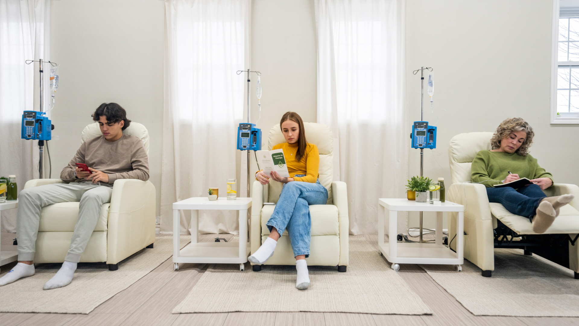 Three people sit in recliner chairs in a bright, modern room, receiving IV therapy from equipment stands beside them.