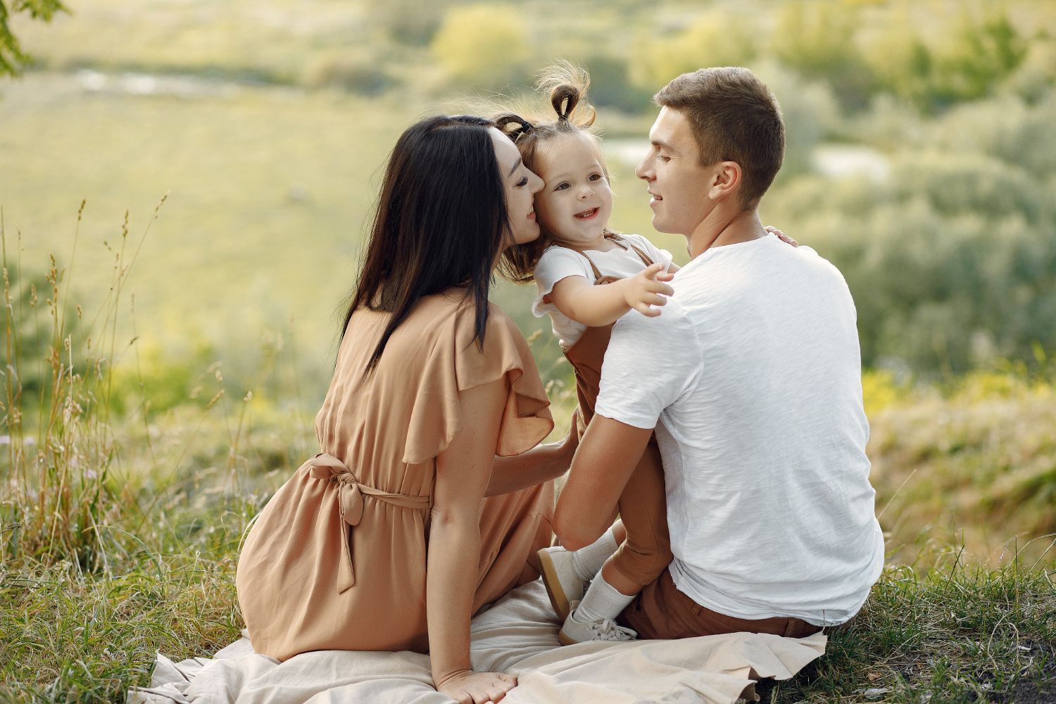 A family of three sitting on a blanket in a field, with parents looking at their laughing child in the center.