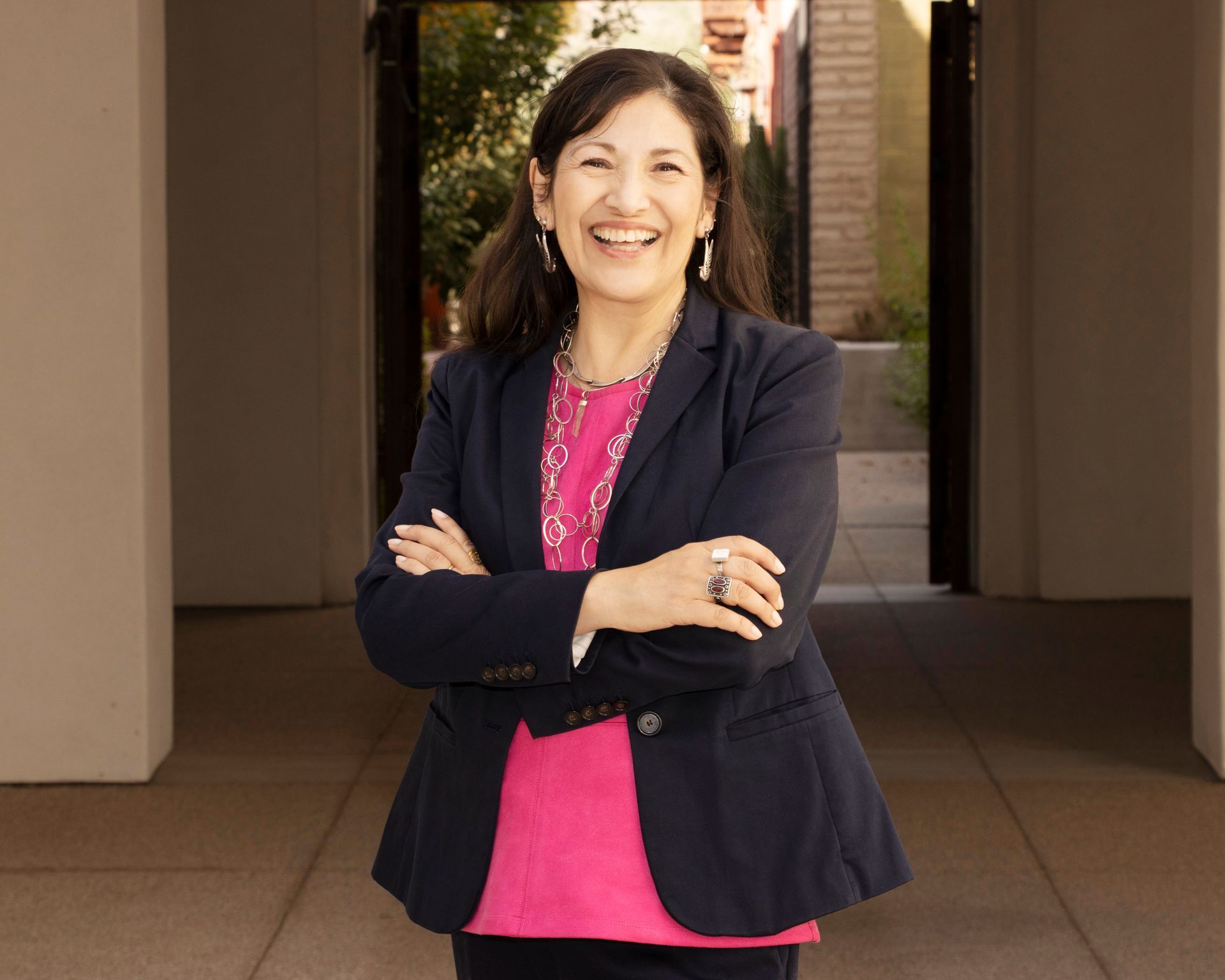 A woman in a blue jacket and pink shirt is standing with her arms crossed and smiling.