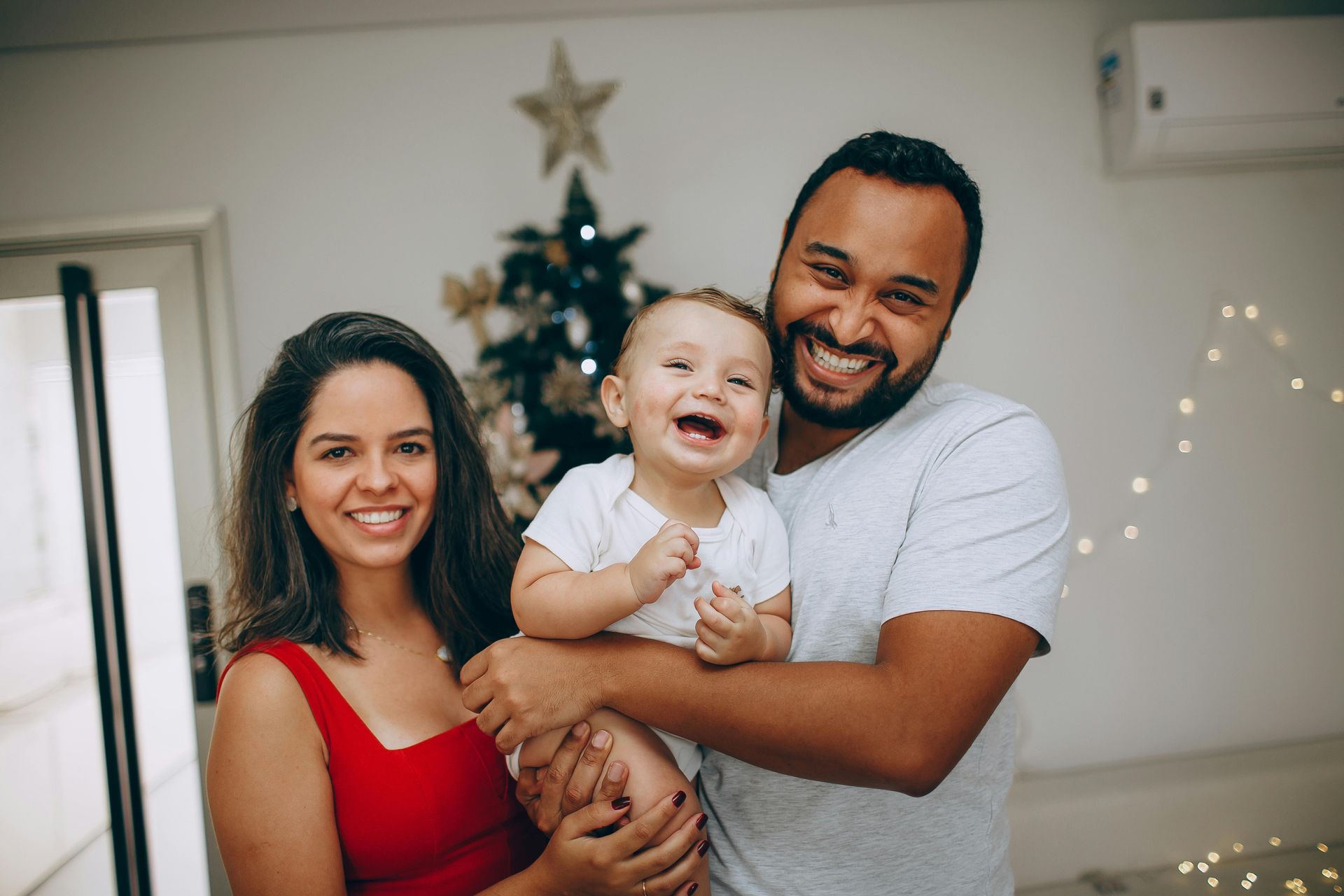 A family of three smiling together indoors with a lit Christmas tree in the background.