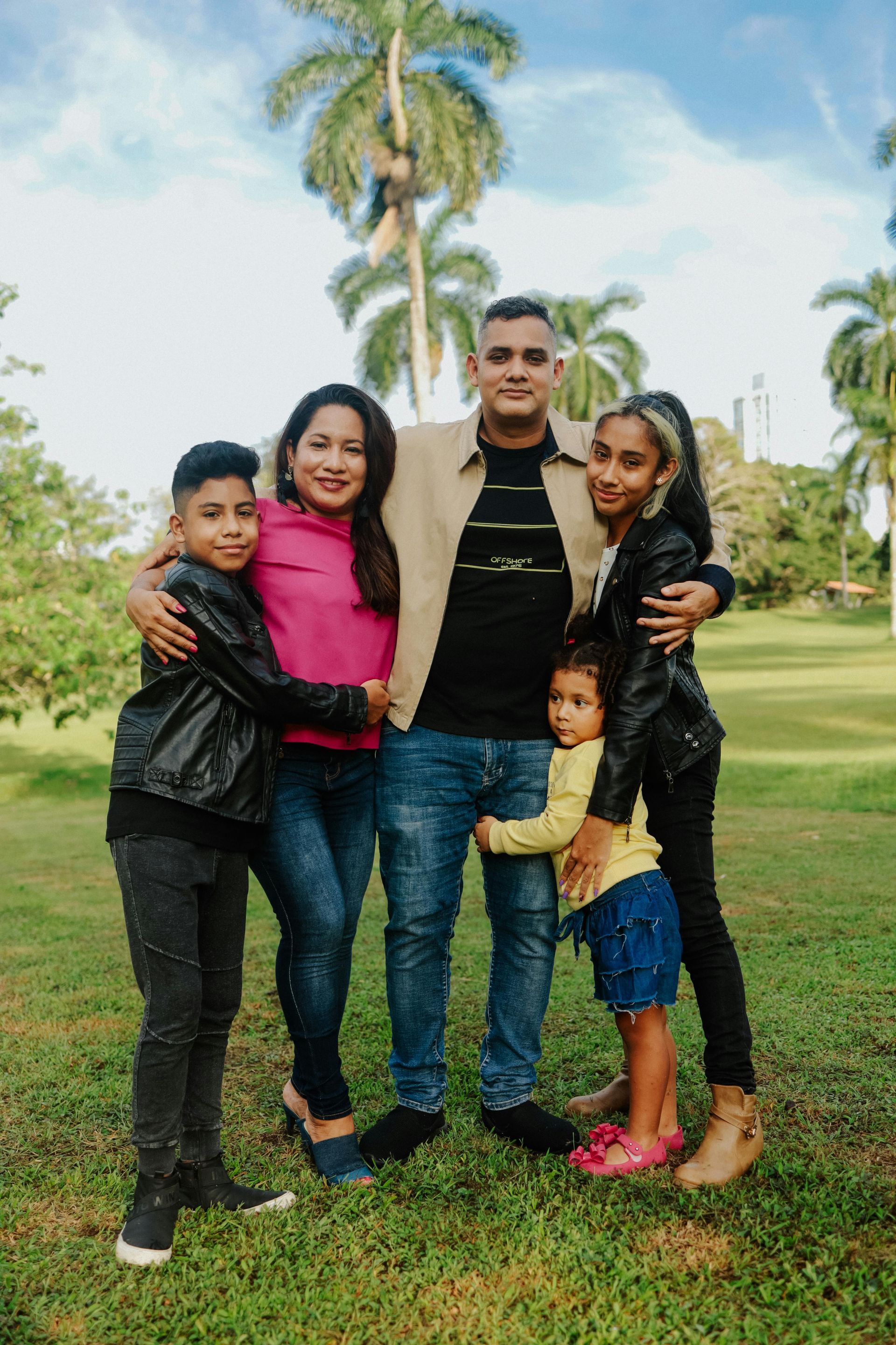 A family stands together in a grassy park with palm trees in the background, smiling as they embrace for a photo.