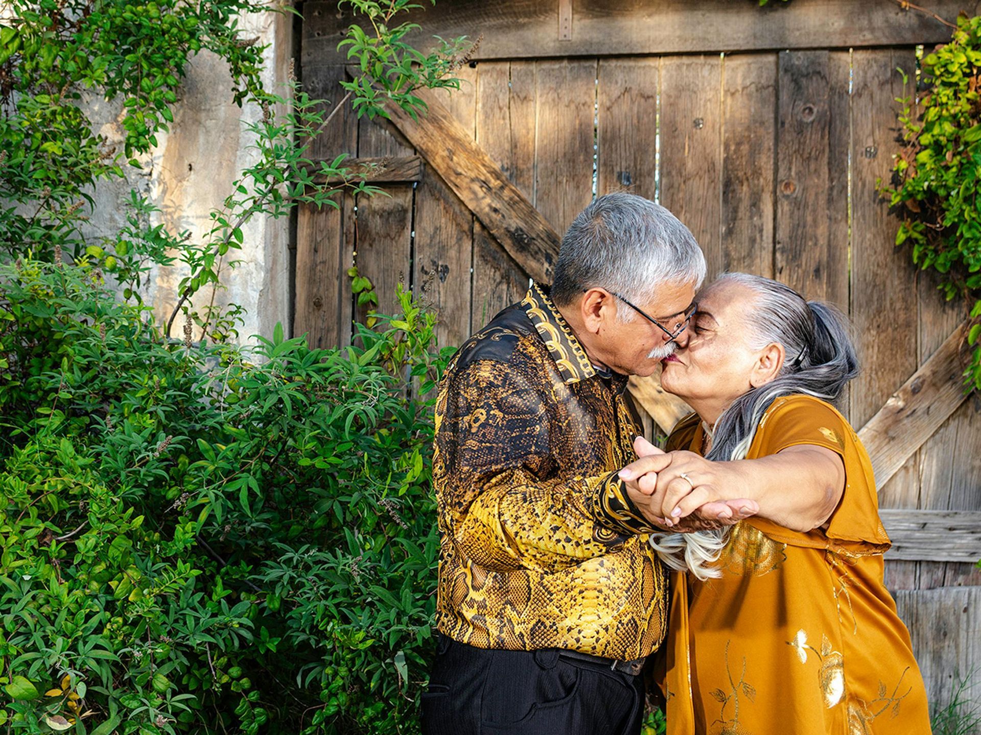 An older couple in gold-toned clothing dancing closely outdoors near a wooden gate and lush green foliage.