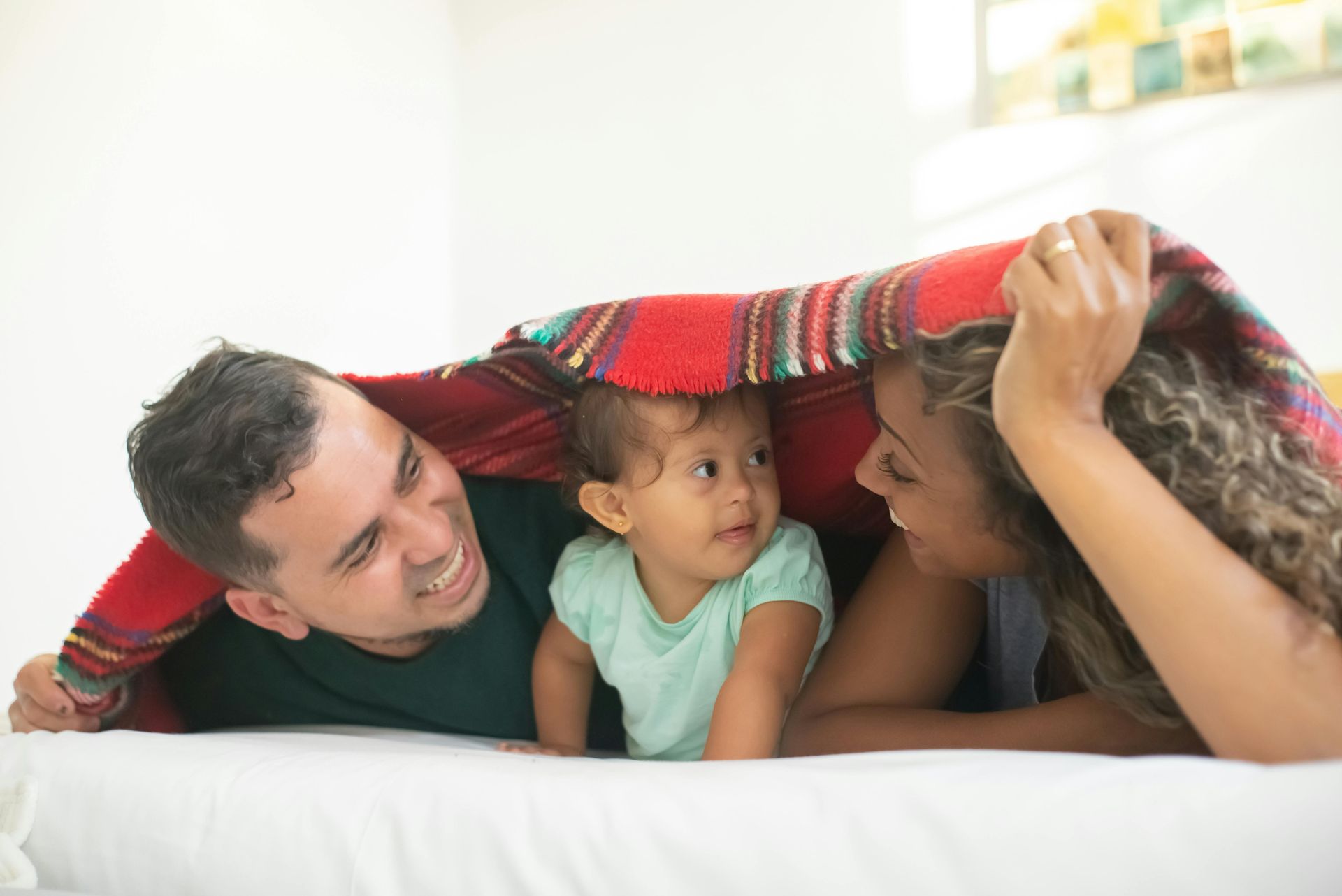 A happy family of three lying together on a bed under a red patterned blanket.