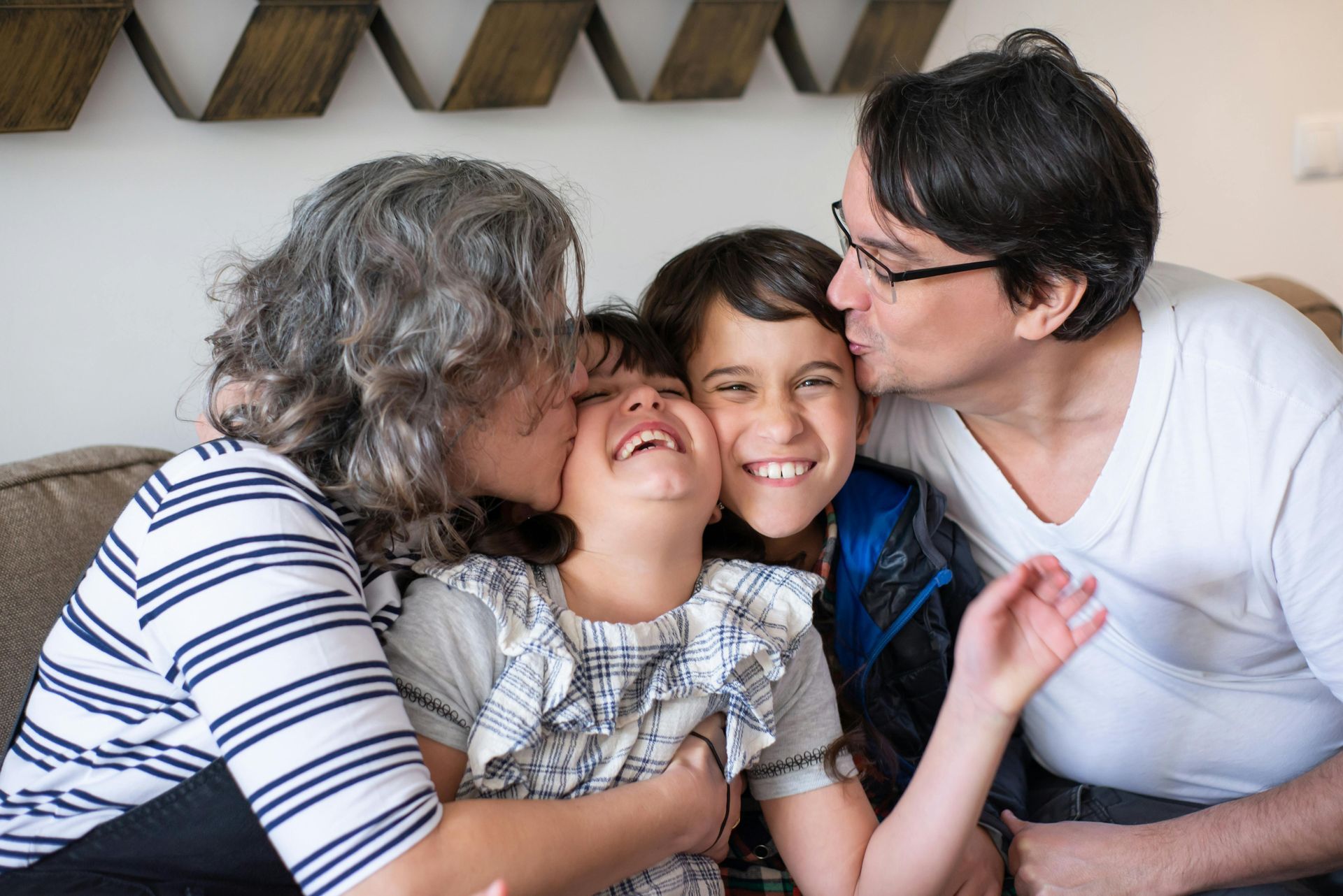 Parents kissing their two children on the cheeks while they laugh together on a sofa.