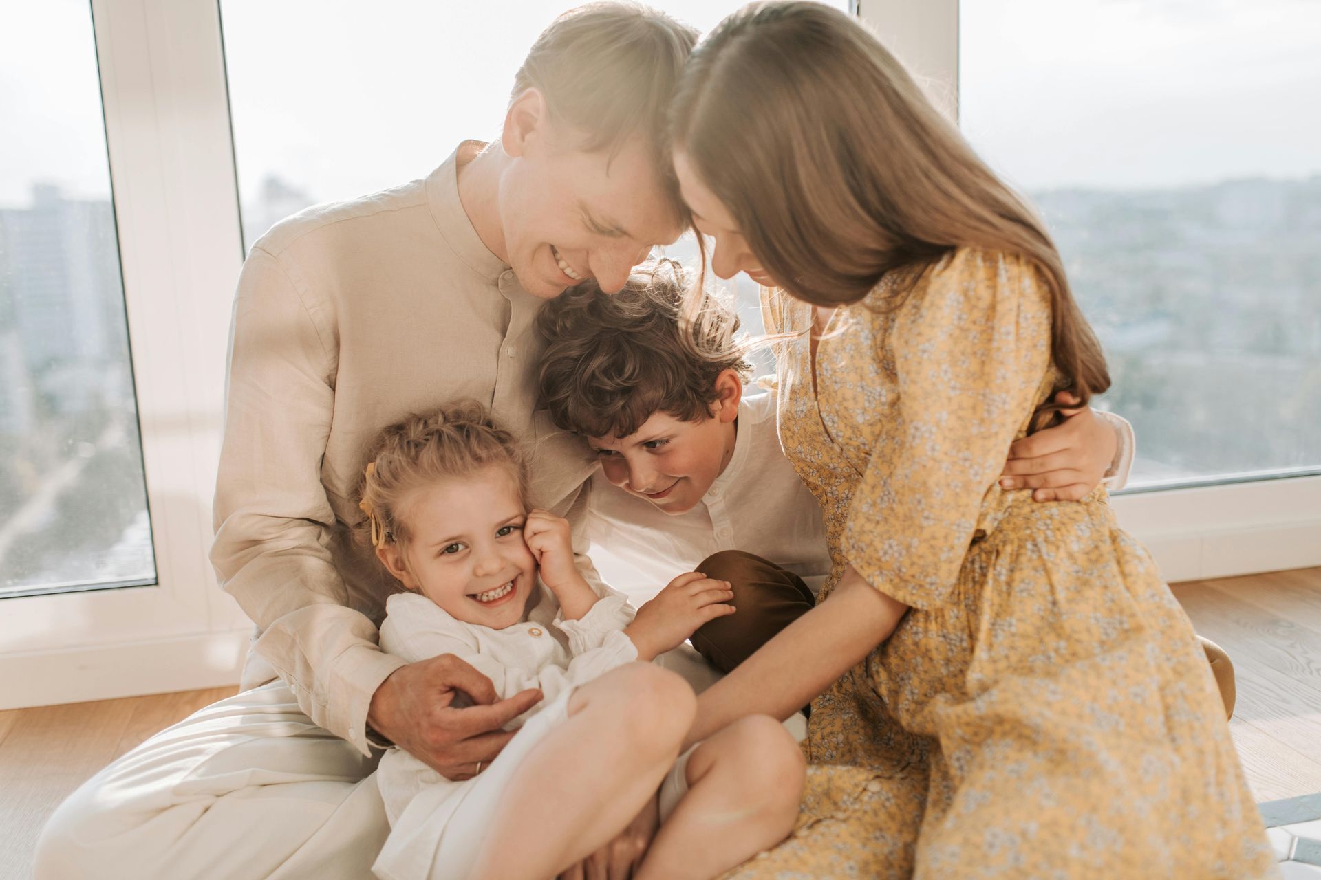 A family of four sits together by a window, smiling and embracing in a sunlit room.