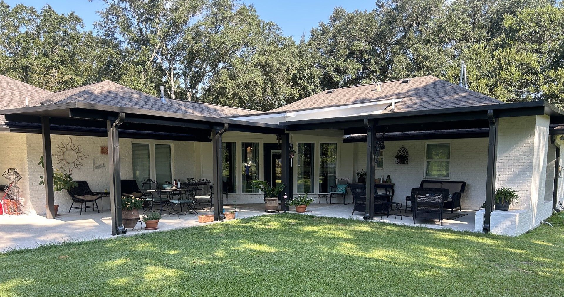 Backyard patio with seating, plants, and black-trimmed roof. Sunny day.
