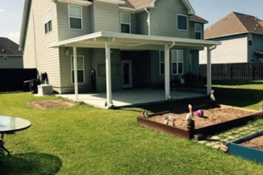 A backyard with a two-story house and covered patio.  There is a sandbox and lawn furniture.