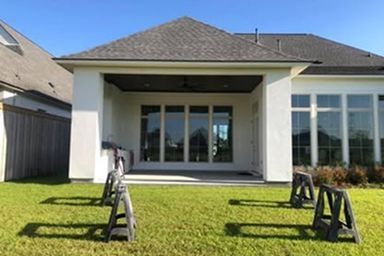 Backyard patio of a white house with large windows and a gray roof, surrounded by green grass; black sawhorses in foreground.