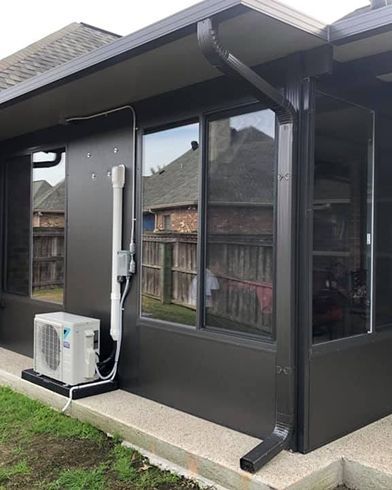 Exterior of a dark-colored building with windows, a gutter, and an air conditioning unit on a concrete base.