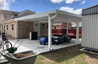 White carport attached to a beige house, over a concrete patio with some lawn equipment.