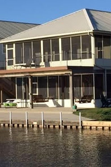 Waterfront house with screened-in porches, white siding, and metal roof. Dock with white posts in foreground.