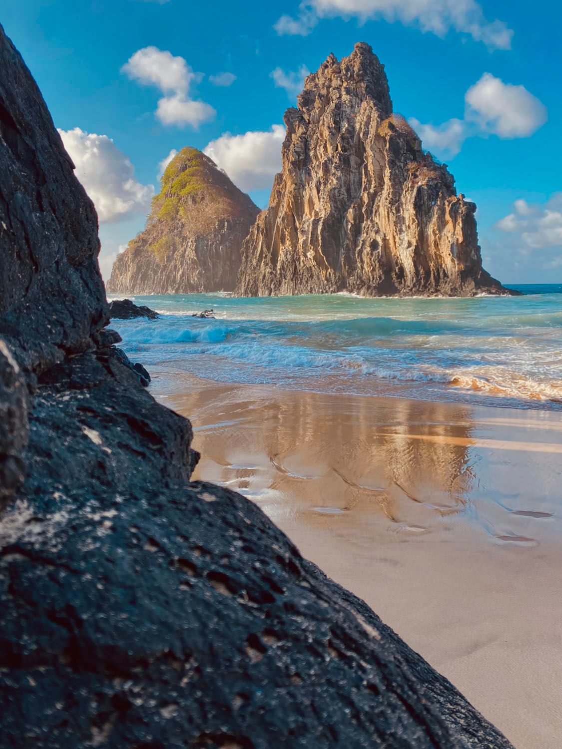Uma praia com uma grande rocha em primeiro plano e uma grande rocha ao fundo.