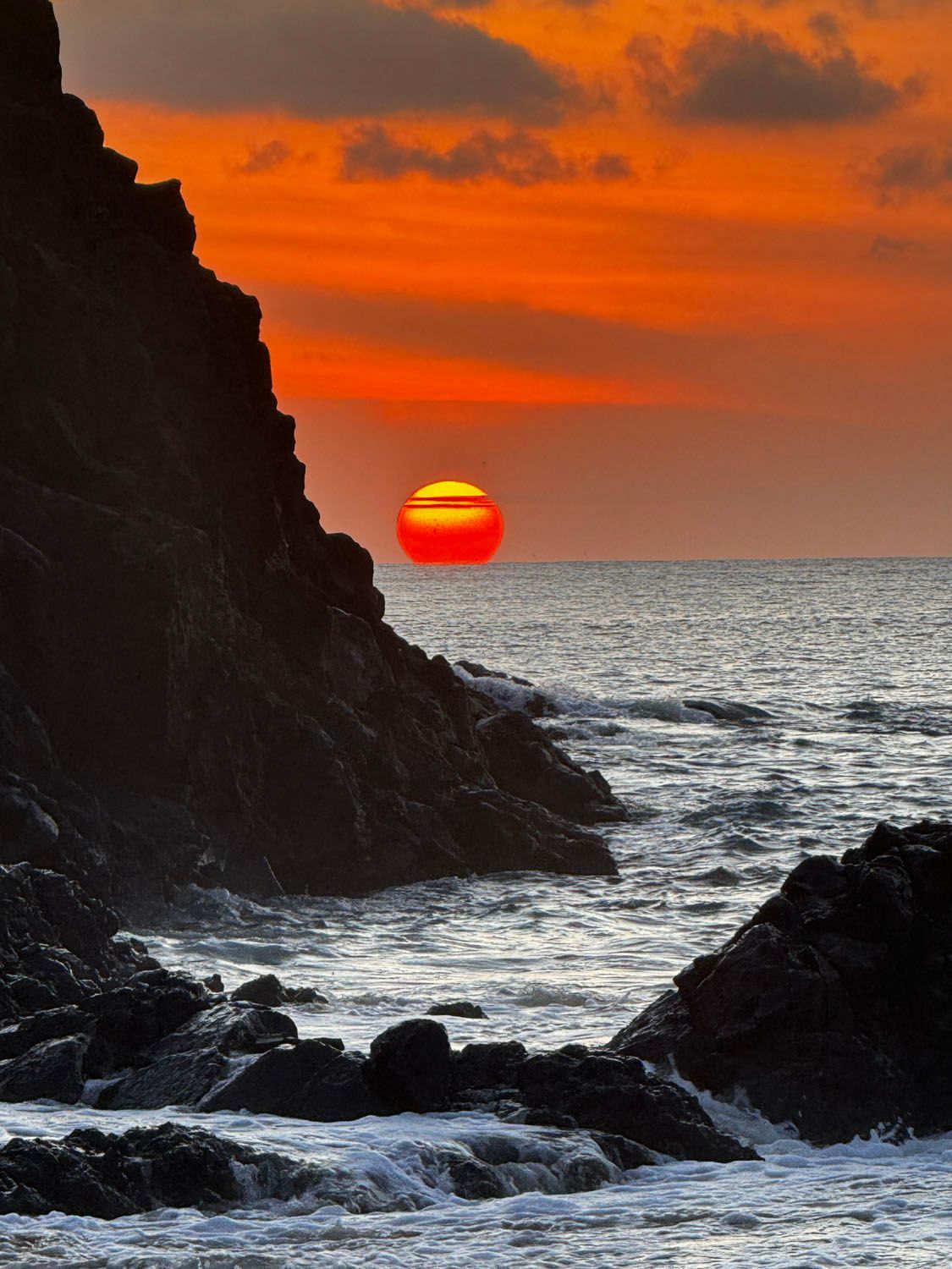 O sol está se pondo sobre o oceano atrás de um penhasco rochoso