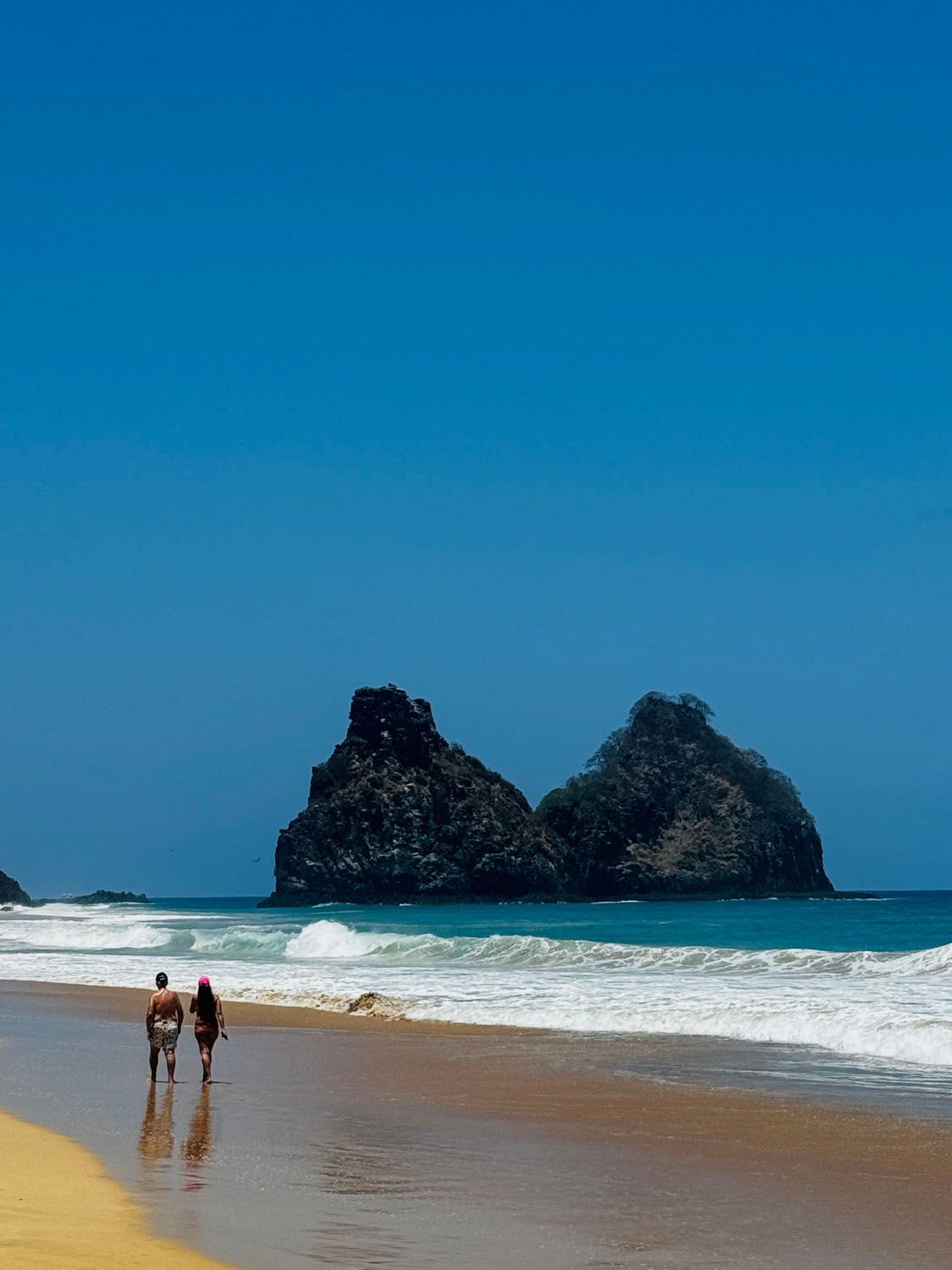 Duas pessoas estão caminhando em uma praia com uma grande rocha ao fundo.