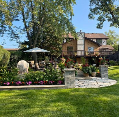 A stunning backyard featuring a stone patio with a dining area under an umbrella, vibrant flower beds, and manicured green grass. Decorative stone columns and lush landscaping create a peaceful outdoor retreat.