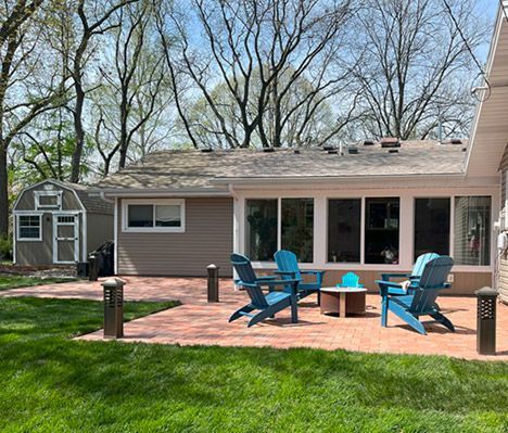 A well-maintained backyard featuring a red brick patio with four blue Adirondack chairs around a small fire pit. The patio is surrounded by lush green grass and illuminated by stylish outdoor lights. A beige house with large sliding glass doors and a storage shed in the background completes the inviting outdoor space