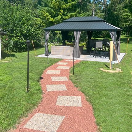 A peaceful backyard setting featuring a gazebo with a black metal roof and curtain panels, surrounded by lush greenery. A decorative stone and gravel pathway leads to the gazebo, creating a welcoming outdoor retreat with seating and a small bar area.