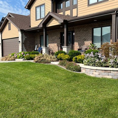 A beautifully landscaped front yard featuring a green lawn, vibrant flower beds, and a raised stone planter. The modern two-story home has a brick and siding exterior with a welcoming front porch, stylish columns, and comfortable seating.
