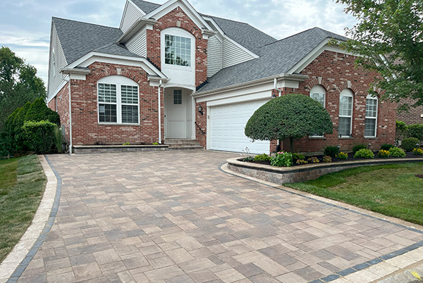 A beautifully designed paver driveway with a custom border leading to a brick home with a white garage. The front yard features a manicured lawn, trimmed bushes, and a raised flower bed, enhancing the home's curb appeal.