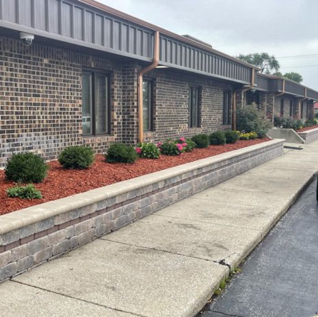A well-designed commercial landscape featuring a long stone retaining wall, neatly trimmed shrubs, and vibrant mulch beds along a modern brick building.
