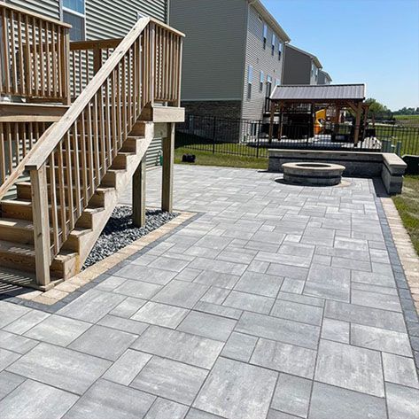 A large backyard featuring a sleek gray paver patio with a built-in fire pit, a wooden deck with stairs, and a covered pergola in the background. The modern hardscape design creates a functional and stylish outdoor living space.