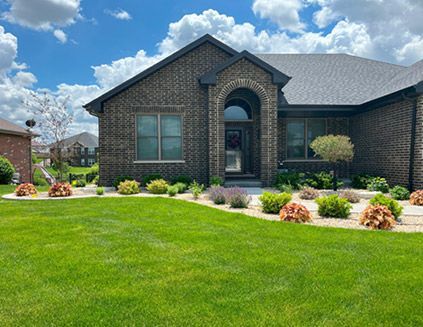 A beautifully landscaped front yard featuring a manicured green lawn, decorative rock beds, and vibrant plants. The modern brick home has an elegant arched entryway and is surrounded by a well-maintained outdoor space under a bright blue sky with fluffy clouds.