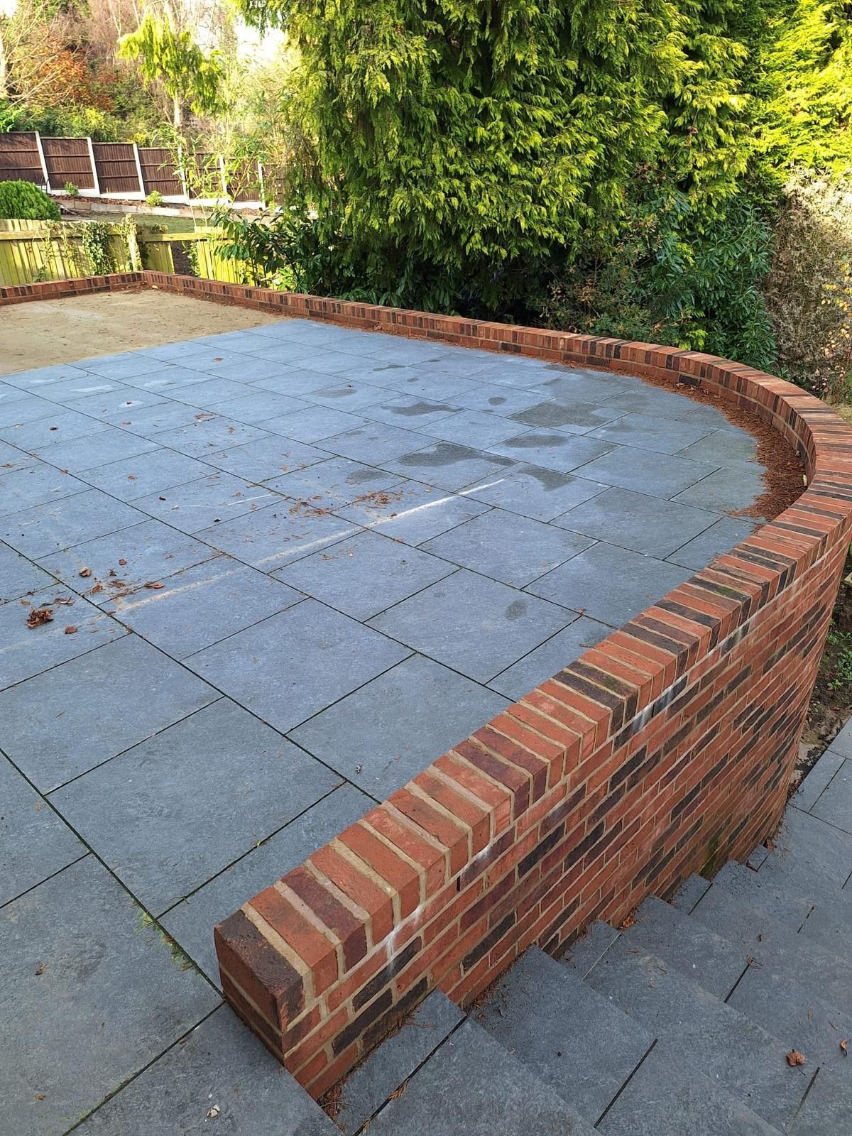Brick wall curves around a paved patio with steps leading down to the right. Trees and water in the background.
