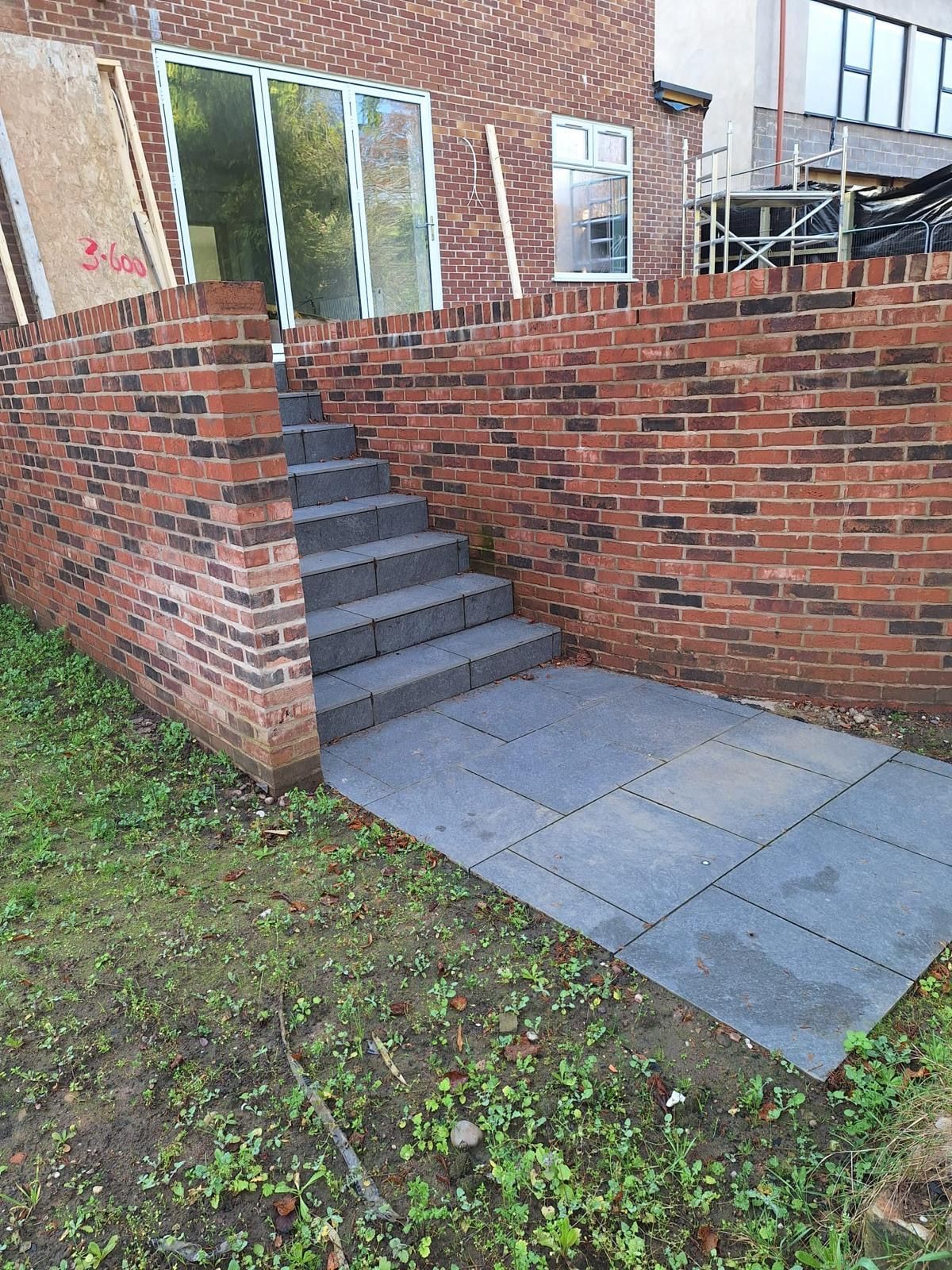 Brick wall with stone steps leading to a patio area, near a house with glass doors and a window.