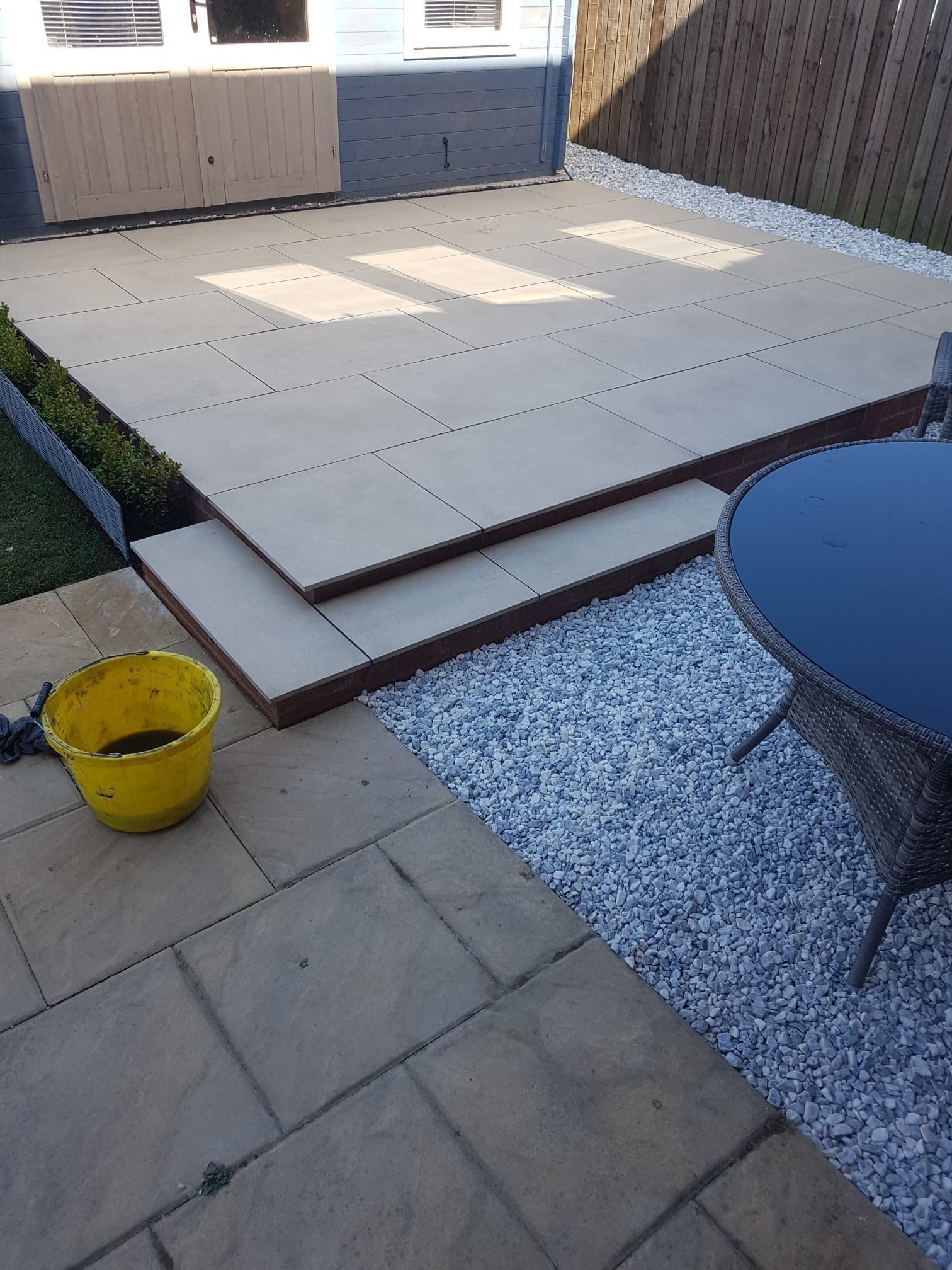 Patio with steps, paving stones, gravel, and a black table. A yellow bucket sits on the patio.