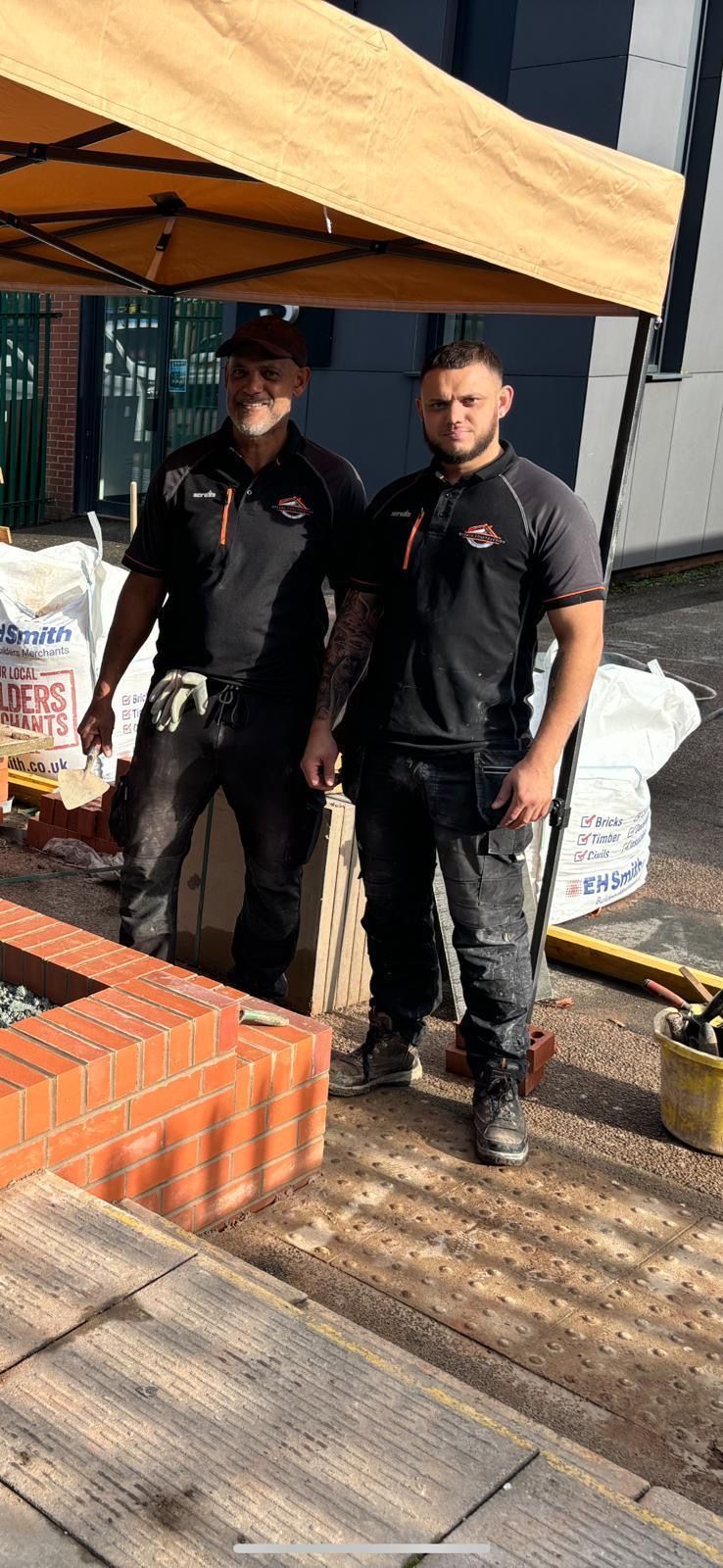 Two construction workers standing by brickwork, under a beige tent.