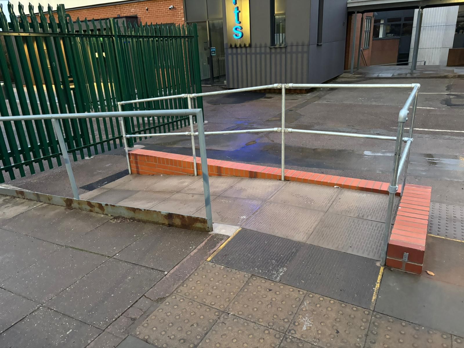 Ramp with metal handrails leading up to a building entrance, bordered by brick and a metal fence.