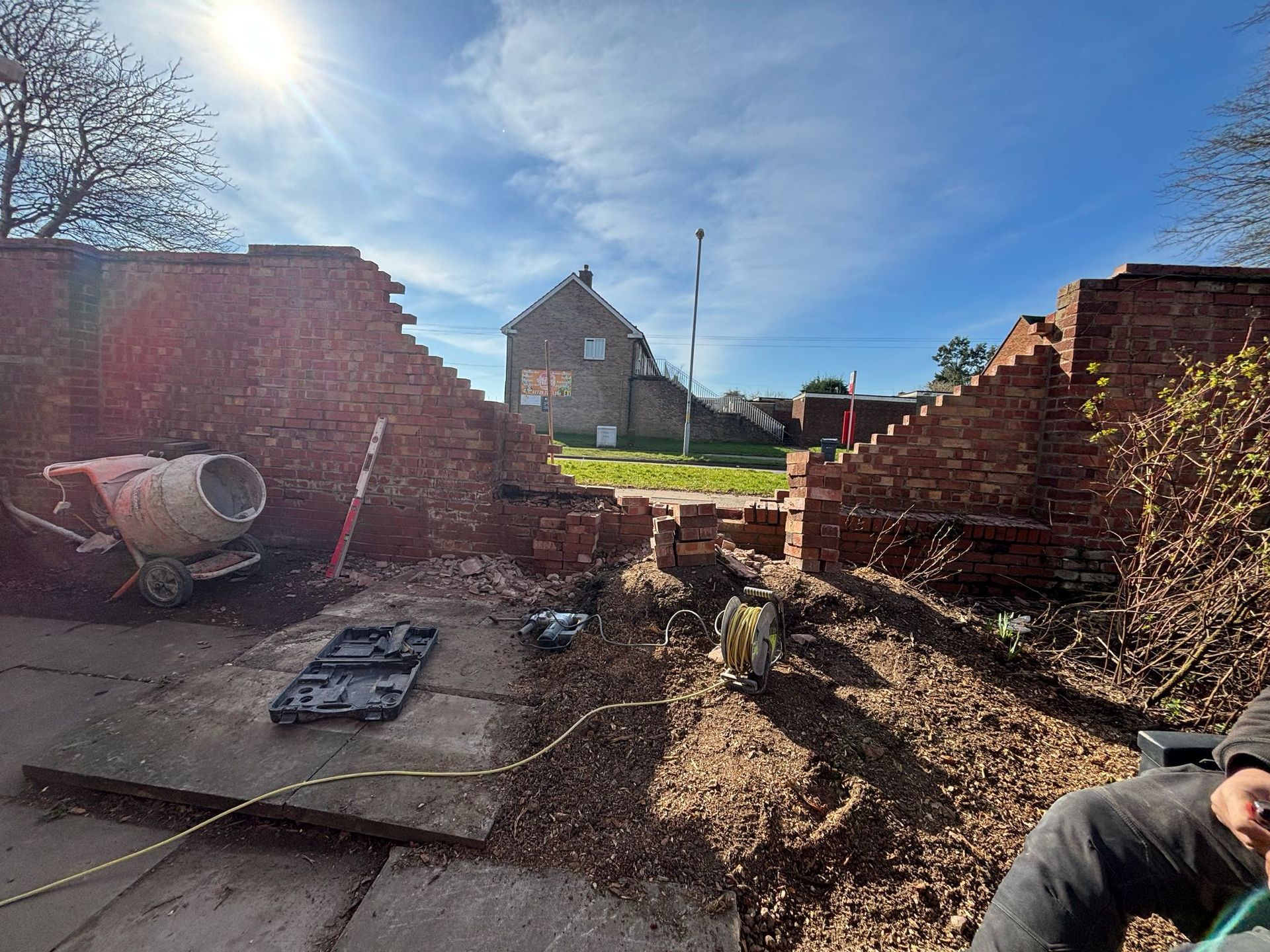 Brick wall under construction, with a concrete mixer and view of a house beyond. Sunny day.