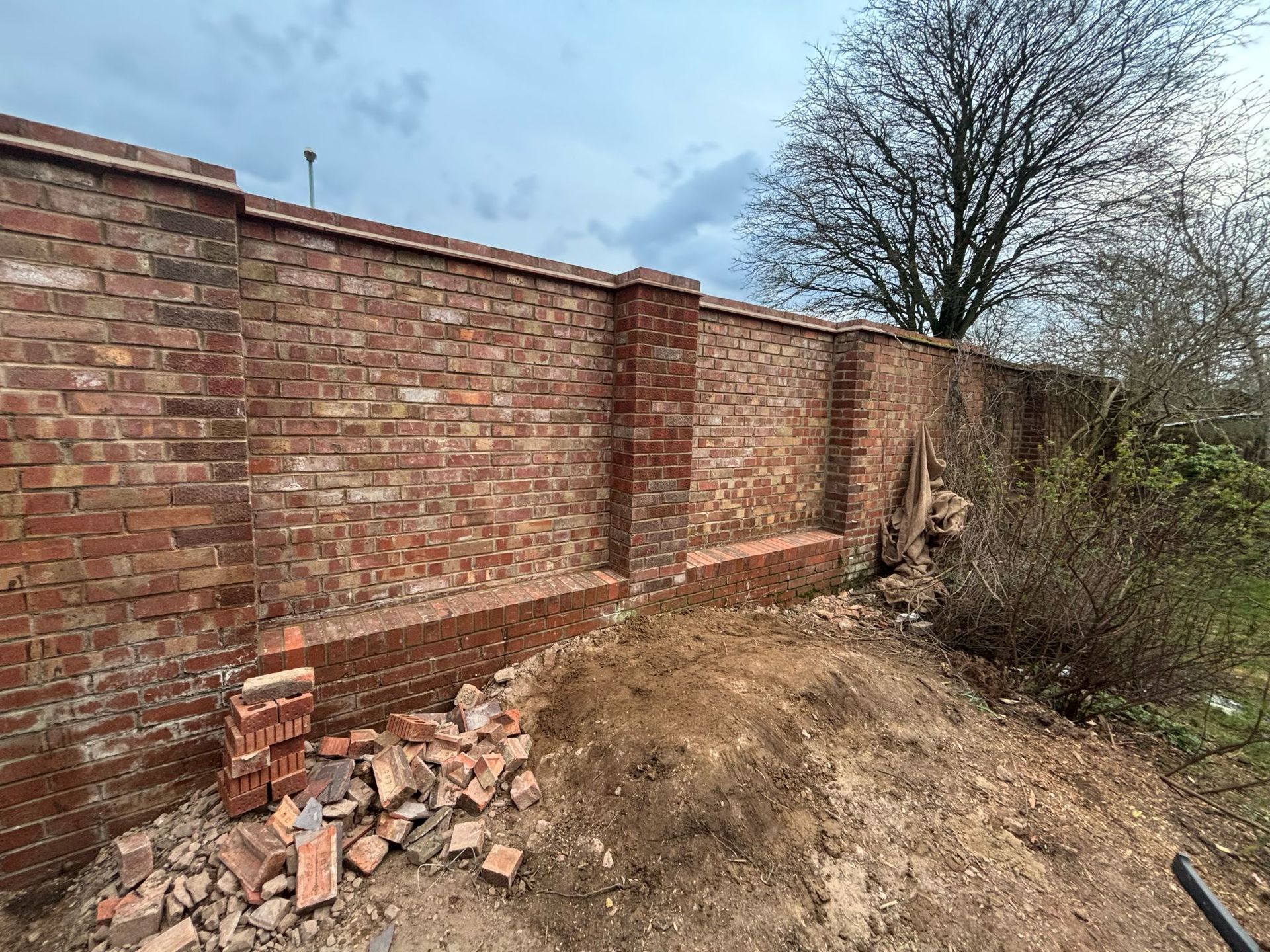 Brick wall under construction, with piles of bricks and soil. Cloudy sky in the background.