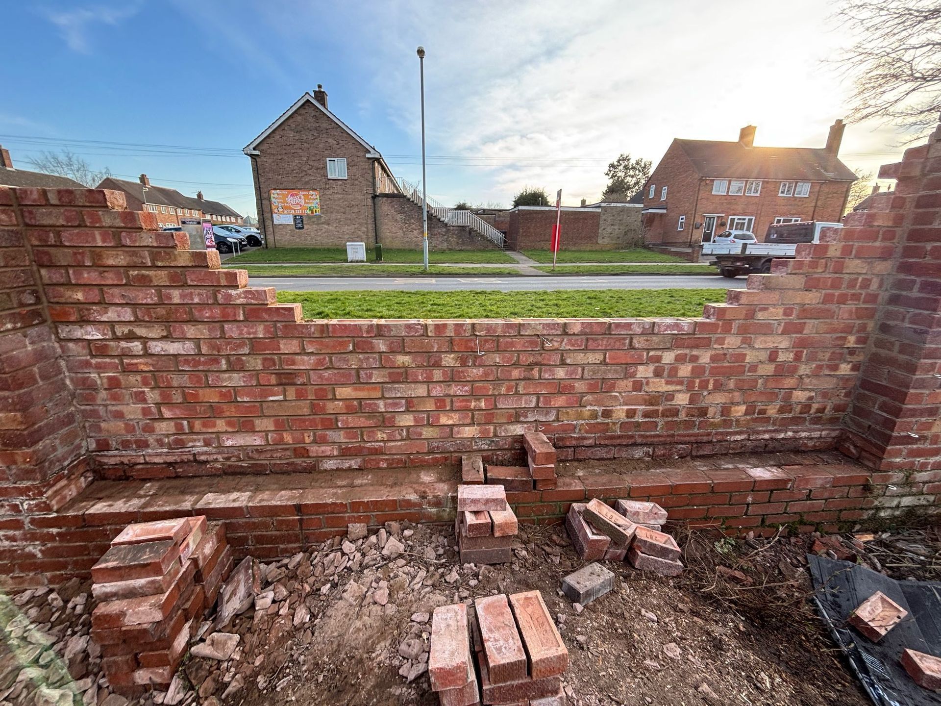 Brick wall under construction, partially demolished with bricks on the ground. Houses and road in background.
