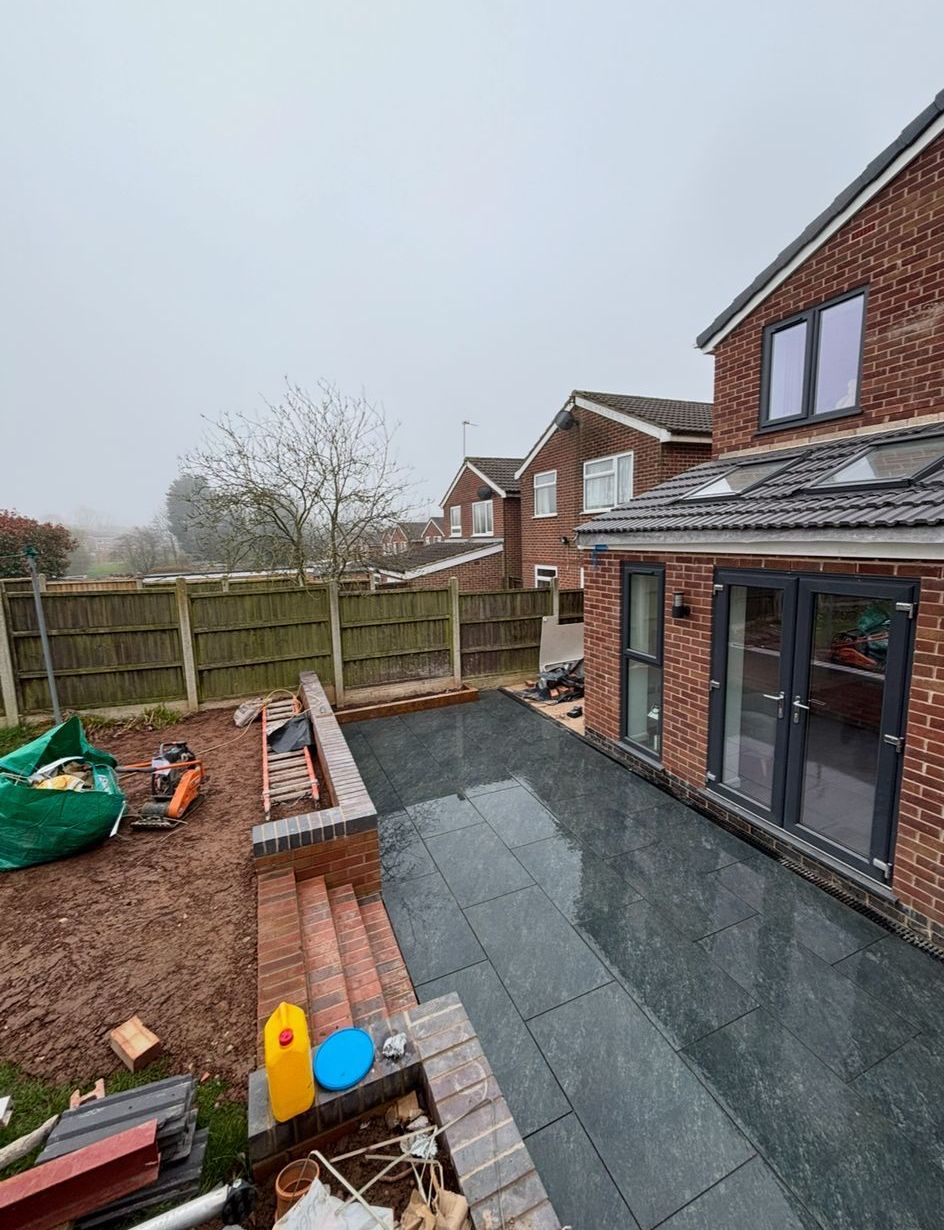 Backyard patio under construction with brick wall, dark grey pavers, and house with glass doors.