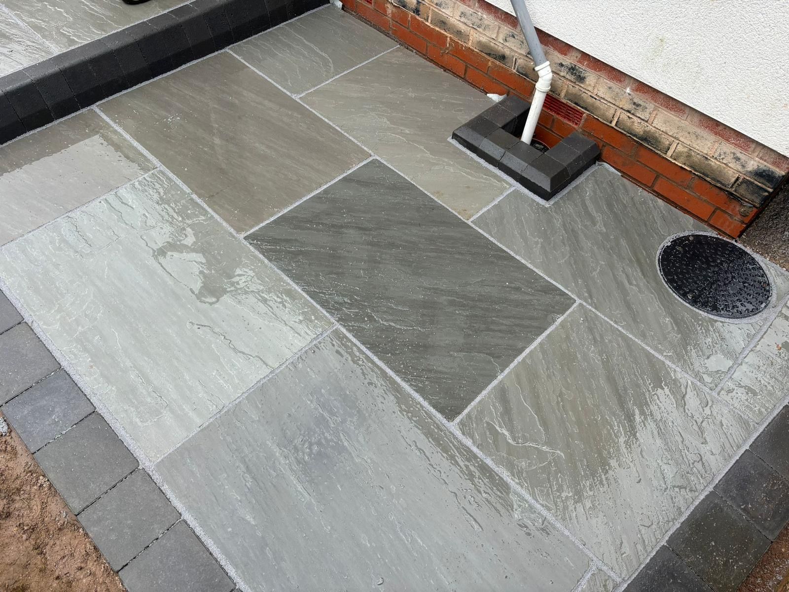 Gray stone patio with dark border and drain, adjacent to a red brick wall.