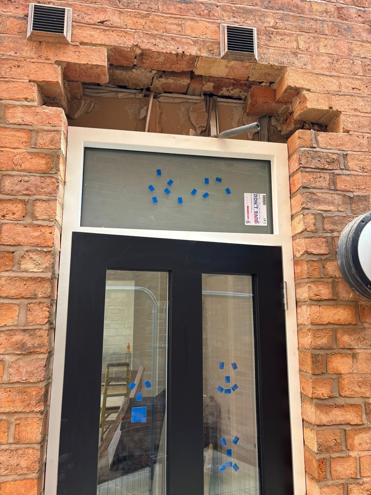 Exterior view of a brick building with a door. The brick above the door is damaged. There are vent covers above.