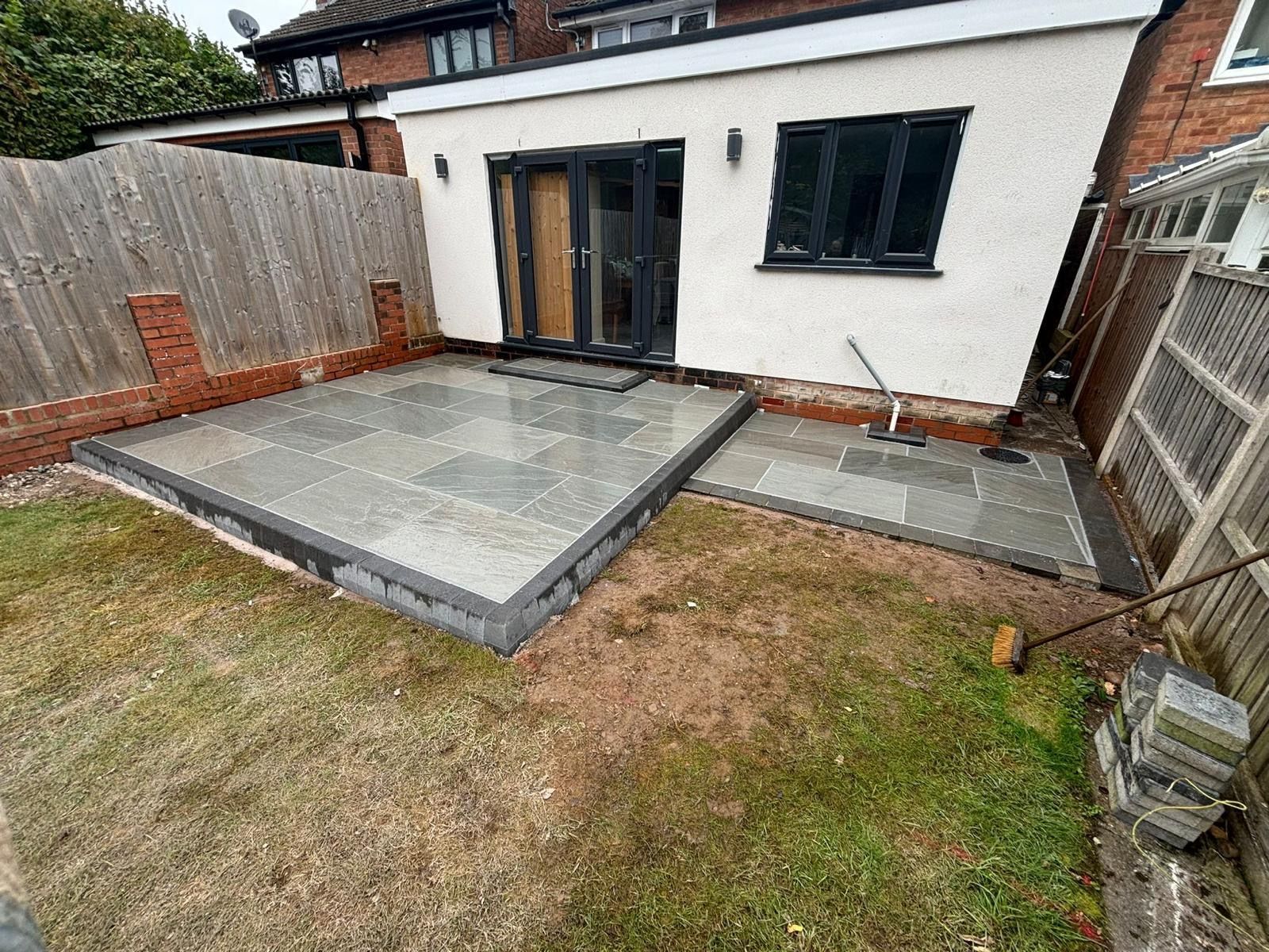 Backyard patio with paved area adjacent to a white building with black framed doors and windows, and a wooden fence.