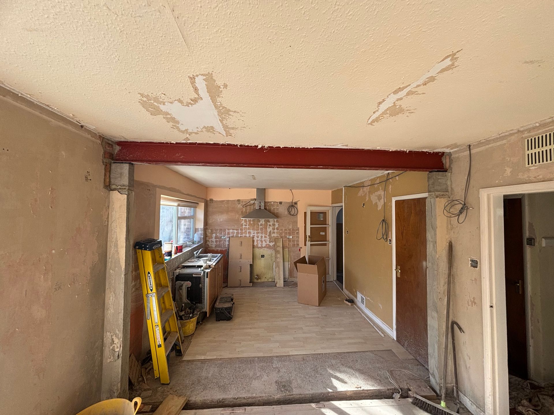 Interior of a room under renovation; red beam, exposed walls, partially removed ceiling, view into kitchen area.