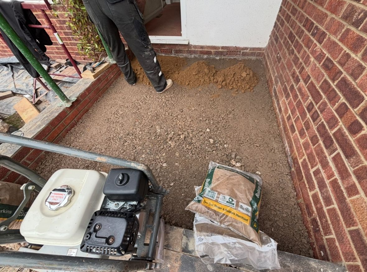 Person compacting gravel with a machine on a patio. Brick wall and bag of sand visible.
