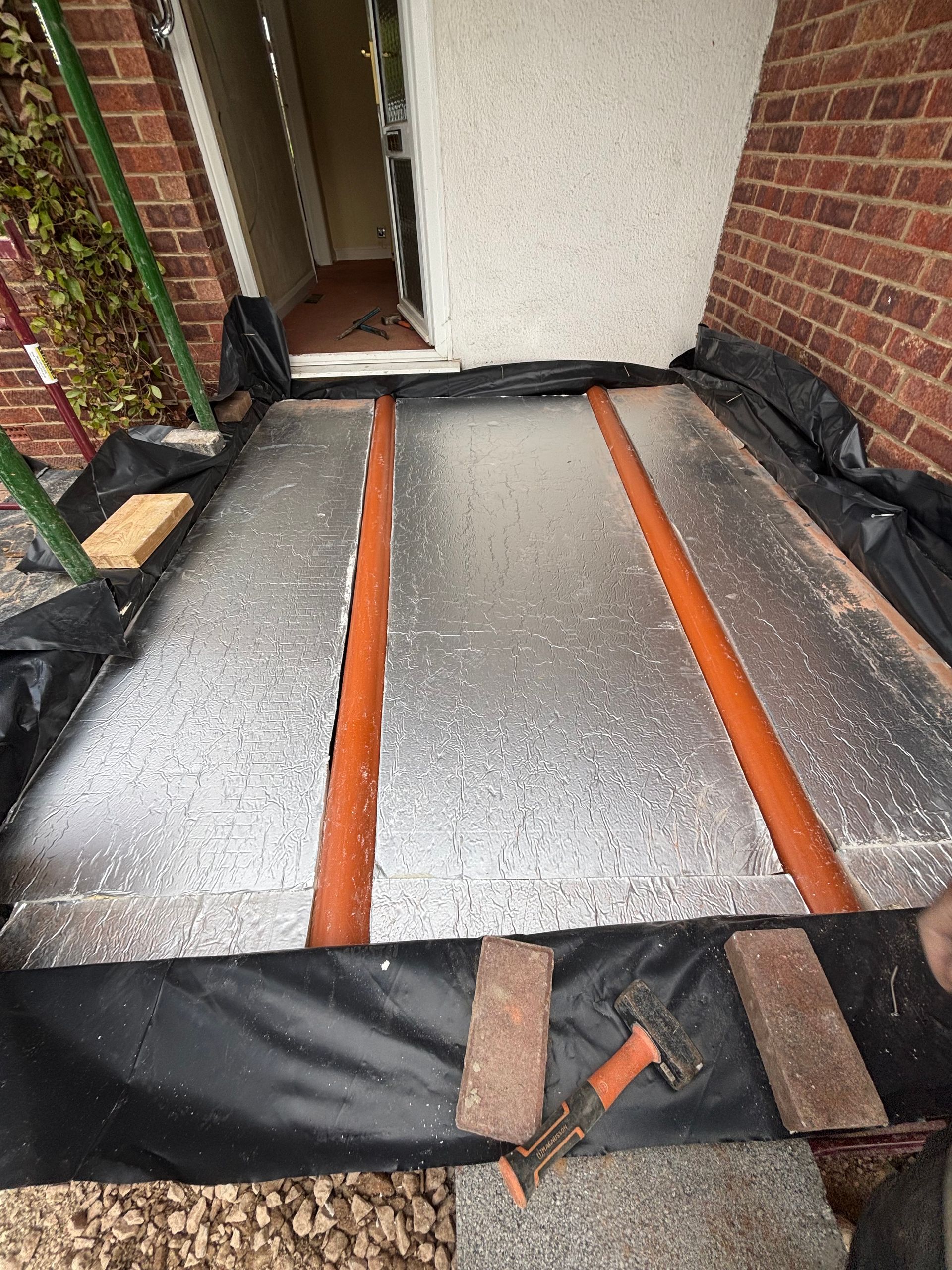 Pathway under construction with insulation, pipes, and protective covering. Brick and doorway in background.
