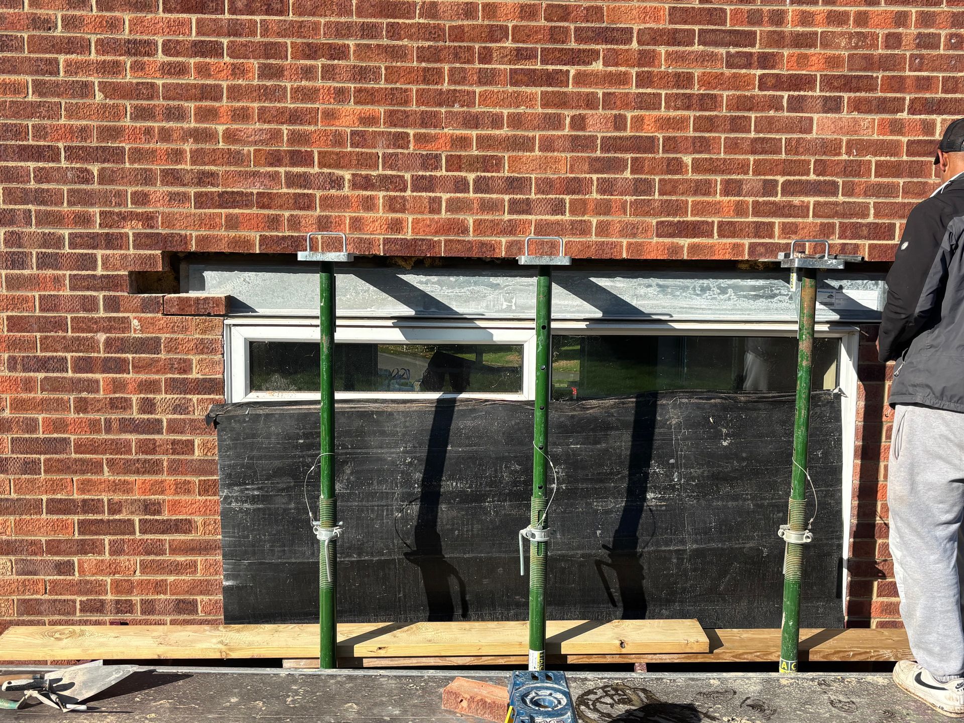 Brick wall with window undergoing renovation; supported by green metal jacks. A person stands nearby.