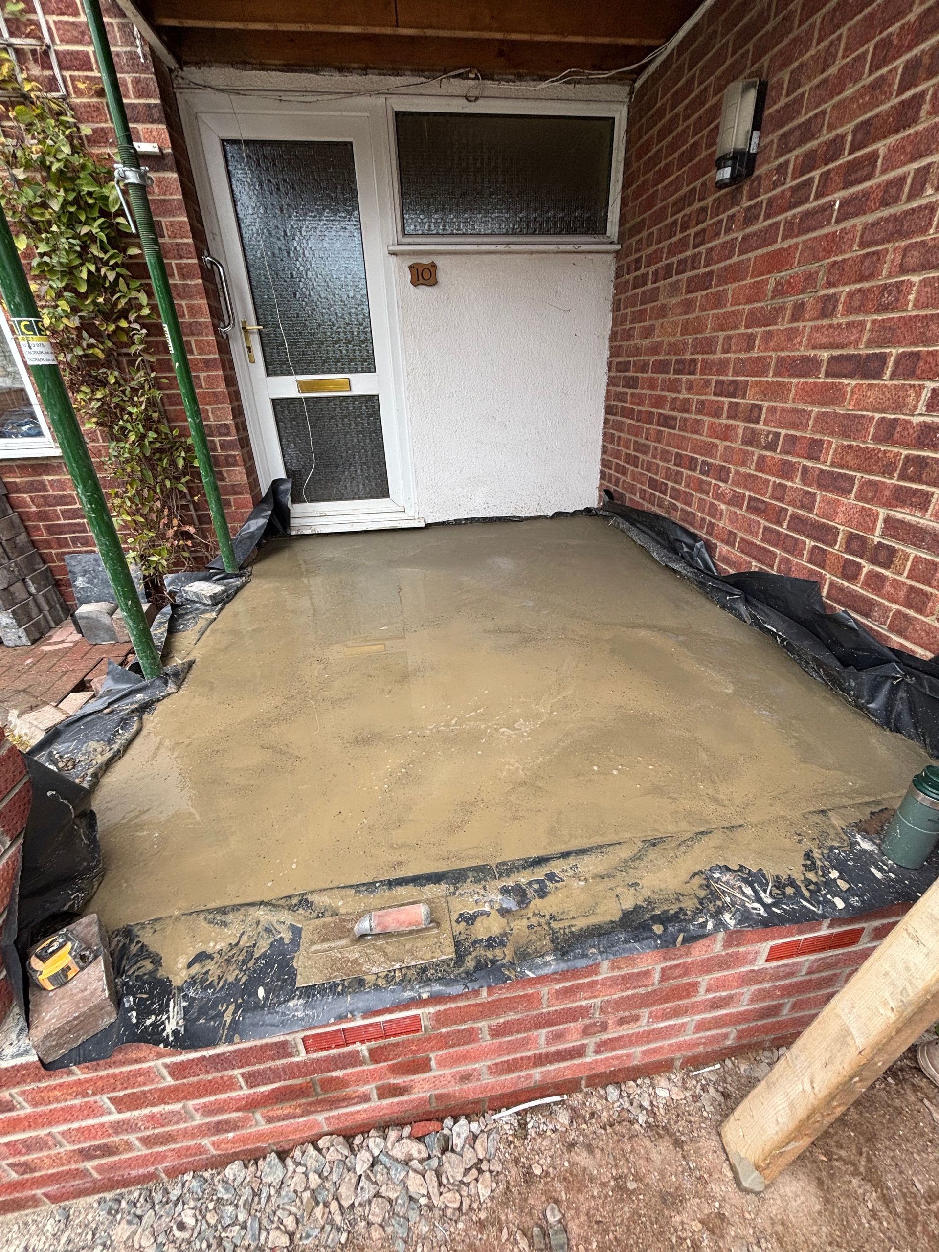 Freshly poured concrete entryway with brick walls and a white door. Black sheeting surrounds.