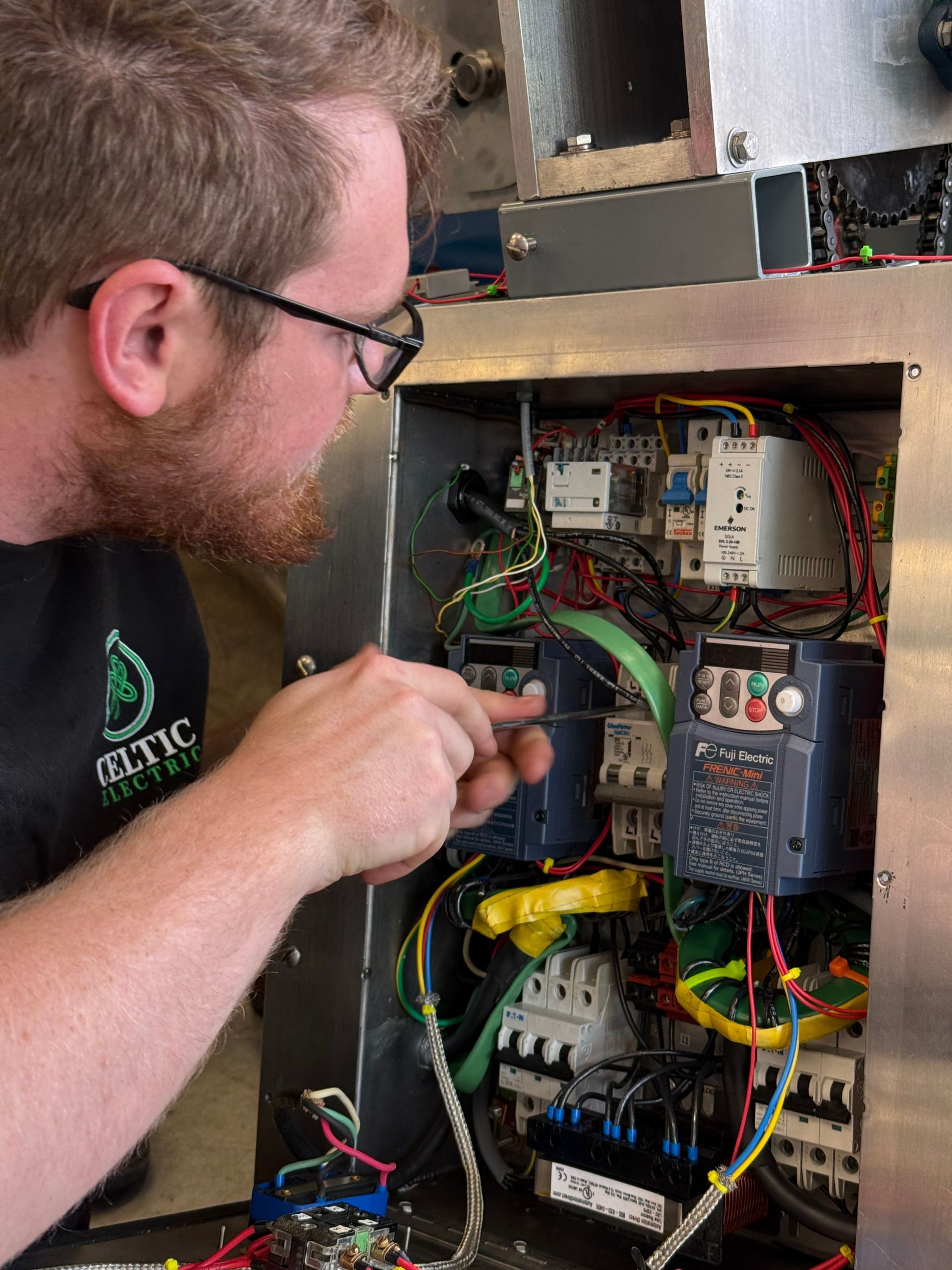Man with glasses inspects an electrical panel, pointing at wiring and components.