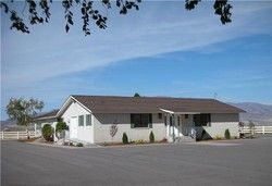 Single-story white building with a brown roof under a blue sky, surrounded by a paved area and a white fence.