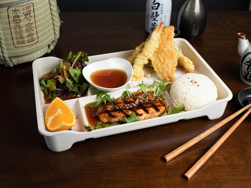 A tray of food with chopsticks and a bottle of sake in the background.